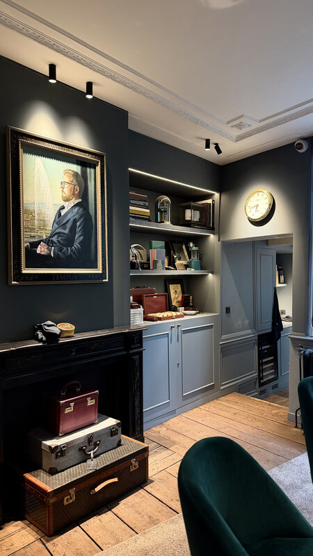 Interior of Amsterdam Vintage Watches — dark walls, framed oil portrait, bookshelves with archival objects, vintage luggage stacked by a fireplace, a green velvet armchair