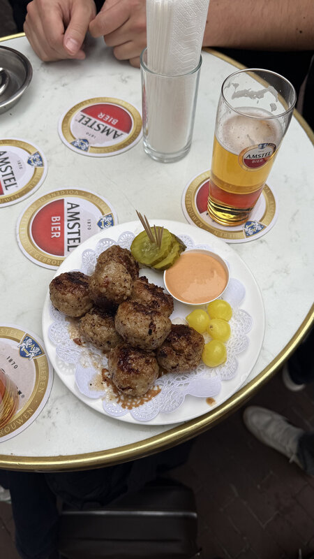 Plate of Dutch bitterballen with mustard, pickles and yellow pickled tomatoes on a table covered in Amstel beer coasters, a glass of Amstel lager alongside