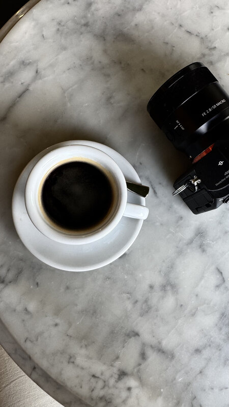 Overhead shot of an espresso in a white cup on a round marble table, next to a Sony FE 2.8 50mm Macro lens