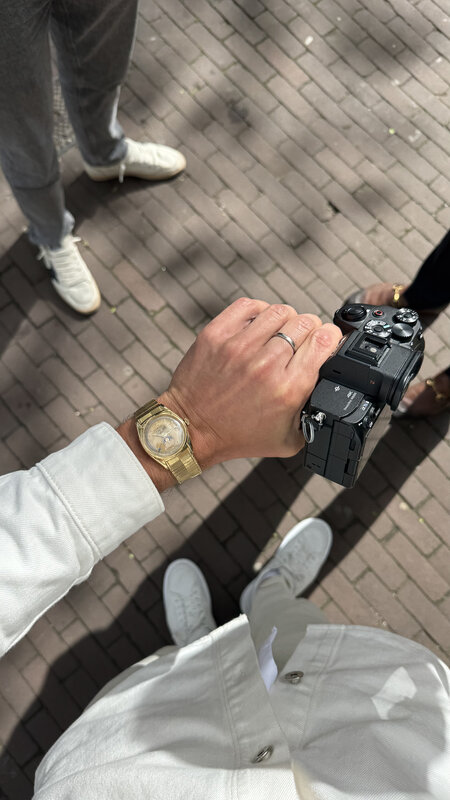 Point-of-view looking down at Jimmy's hand holding a Sony Alpha camera body on a brick Amsterdam street, the gold Rolex Stelline visible on the wrist, white sneakers in frame