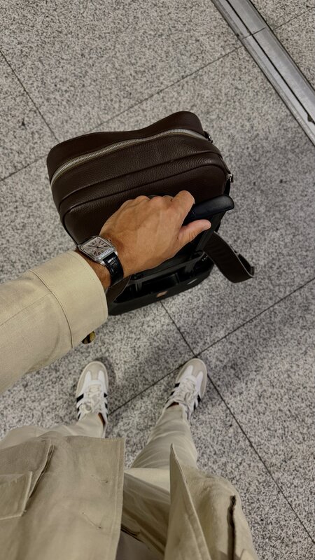 Overhead point-of-view of a traveller walking with a rolling suitcase and a brown leather bag, a steel Cartier Santos on the wrist, on a stone terminal floor