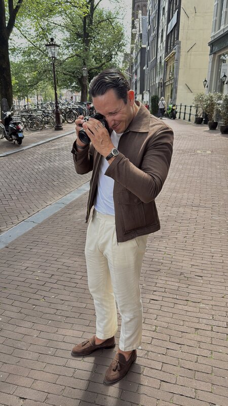 Jimmy Heneidy shooting with a compact camera on an Amsterdam cobbled street, brown jacket and cream trousers, bikes parked along the canal behind him