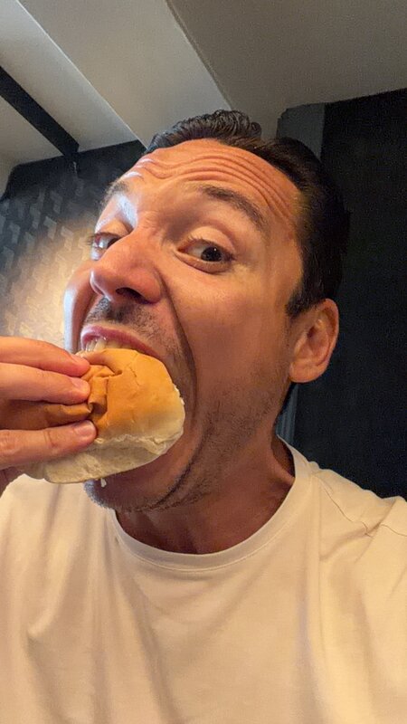 Close selfie of Jimmy taking a bite of a small burger in a dark-walled room