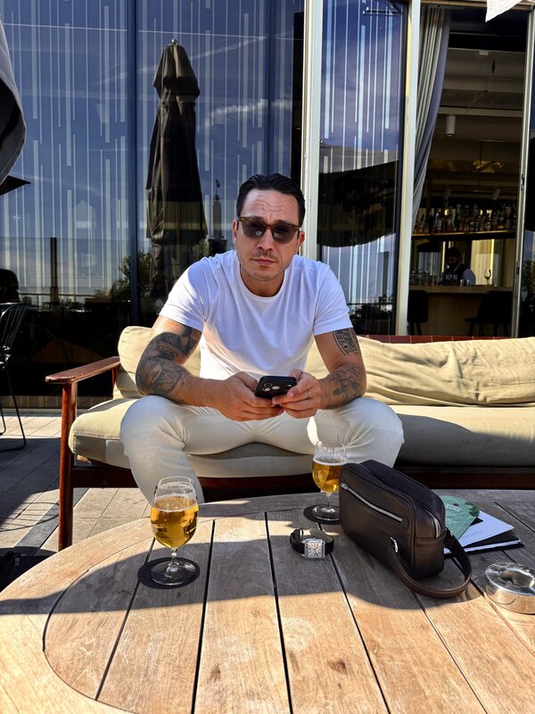 Jimmy sitting on a rooftop terrace sofa at the W Amsterdam, beers on the low wooden table beside a Cartier watch and a leather wash bag