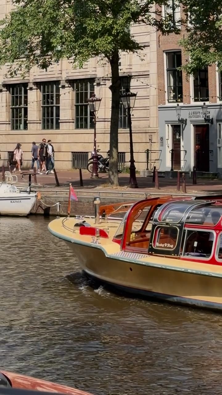 A classic wooden salon boat gliding along an Amsterdam canal in summer sunlight, people walking the quay behind it