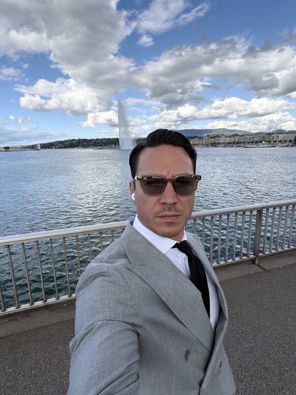 Jimmy Heneidy in a grey suit and dark sunglasses on the lakeside promenade in Geneva, the Jet d'Eau and the far shore behind him