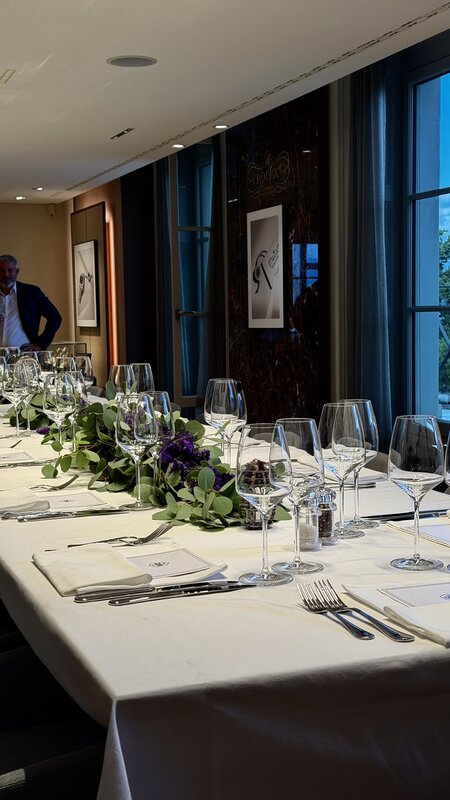 A long dinner table set beside tall windows, with white linen, rows of glassware and low greenery down the centre