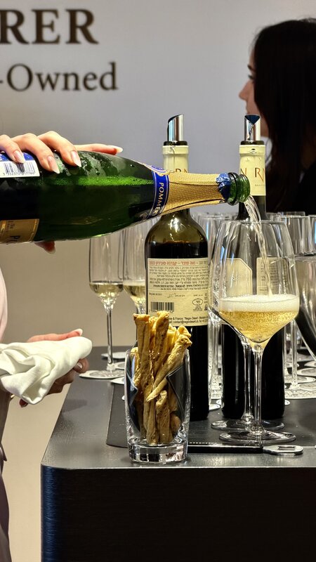 Champagne being poured into a wine glass beside bottles and rows of glassware, with Bucherer Certified Pre-Owned signage behind