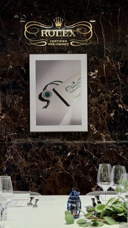 Gold Rolex Certified Pre-Owned signage on a dark marble wall above a framed image of a CPO tag and guarantee card, with the dinner table below