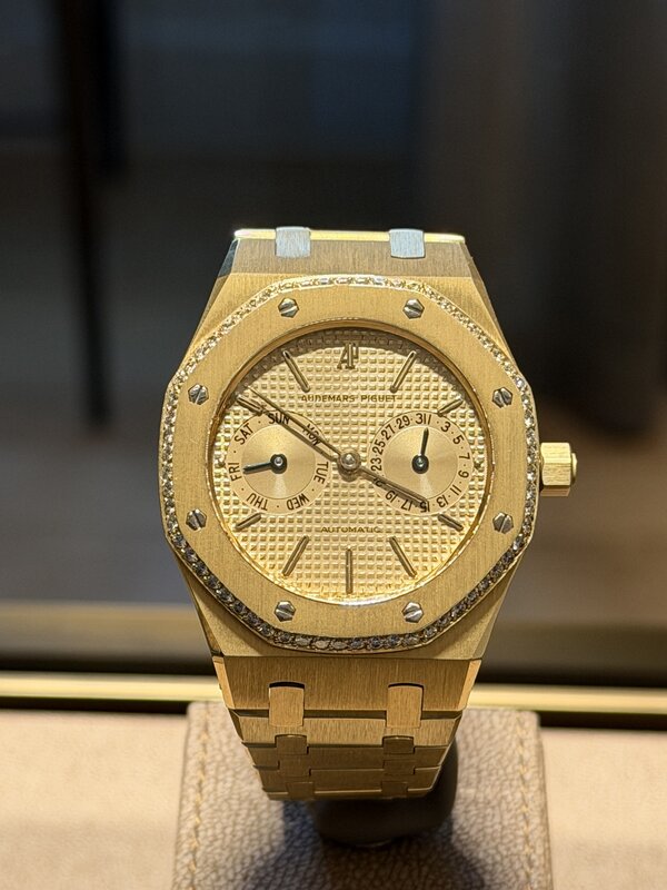A gold Audemars Piguet Royal Oak with a stone-set bezel, day and date subdials and a textured dial, displayed on a cushion behind glass