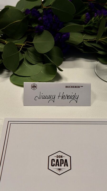A place card reading Jimmy Heneidy on the table beside a Don Capa card and green foliage