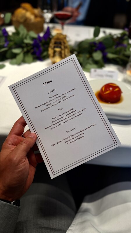A printed three-course menu held above the dinner table, with the tomato starter on a plate behind it