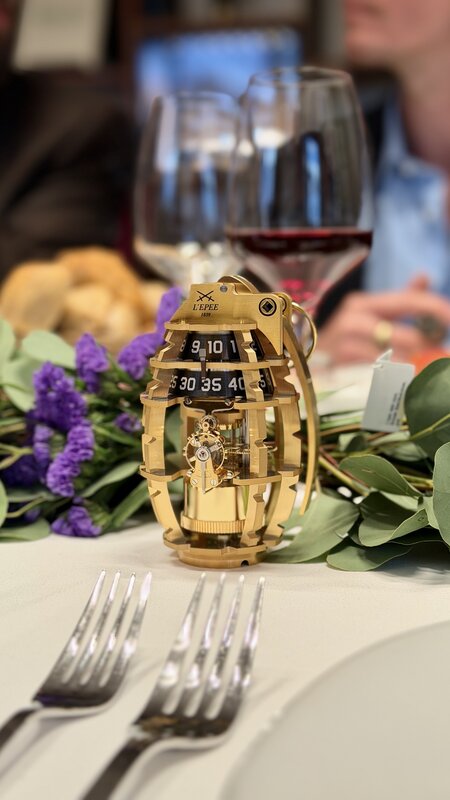 A gold-coloured, grenade-shaped L'Epée 1839 table clock with two numbered drums, standing among the table greenery between forks and wine glasses