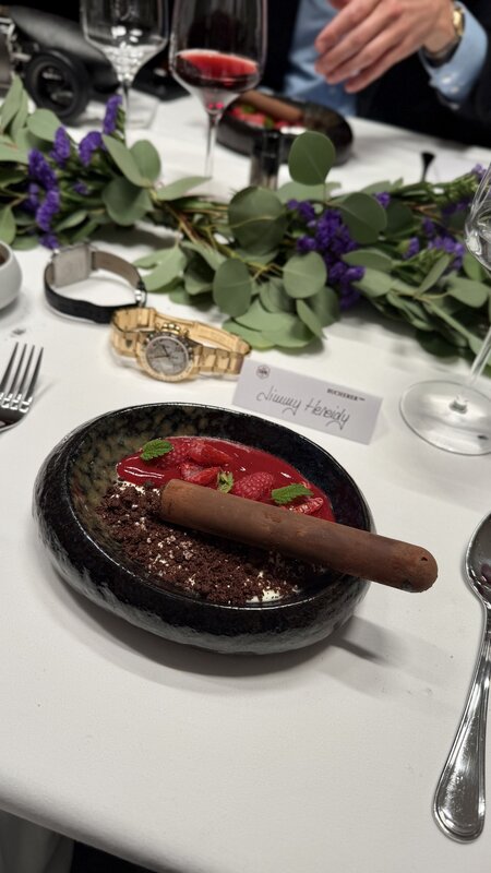 A cigar-shaped chocolate dessert with raspberries and chocolate crumble in a dark bowl, with two watches and Jimmy Heneidy's place card behind it