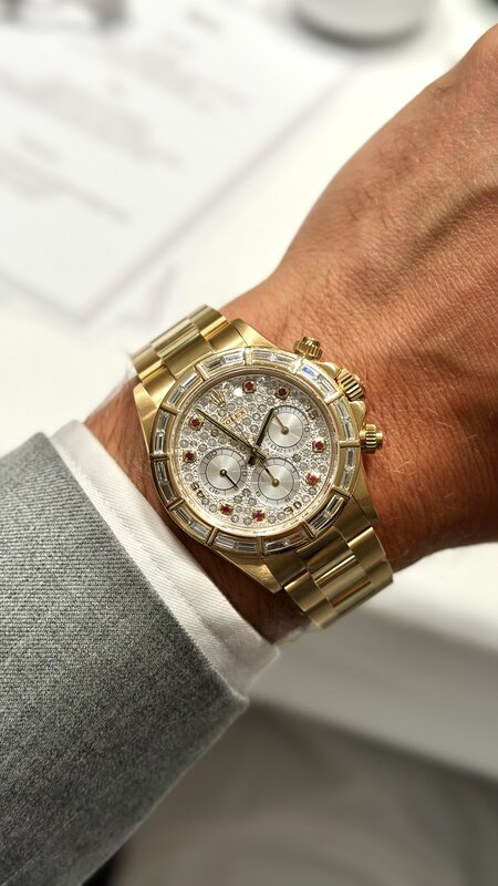 A gold Rolex Daytona with a stone-set dial, red gemstone hour markers and a baguette-cut stone bezel, worn on a wrist above the dinner table