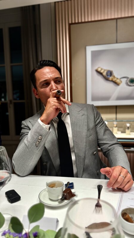 Jimmy Heneidy in a grey suit drawing on a cigar at the table, an espresso cup in front of him, watch displays on the wall behind