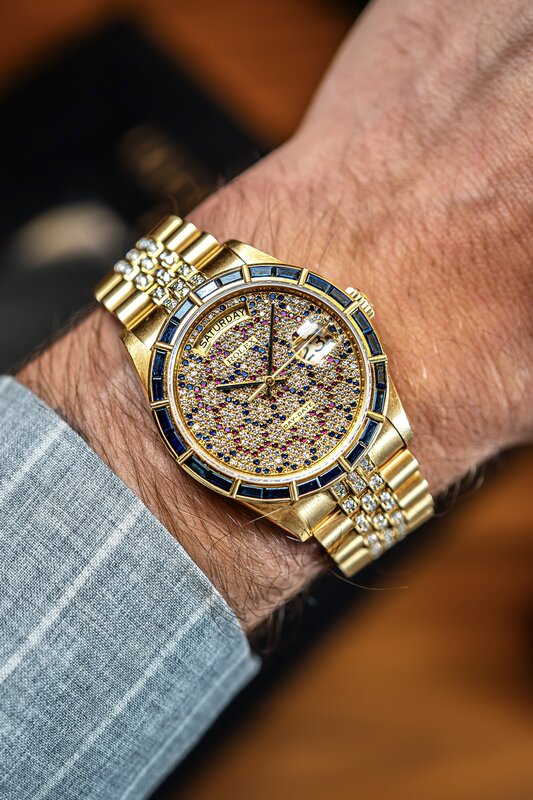 Rolex Day-Date in yellow gold with pavé diamond dial and rainbow gemstone bezel on wrist, blue striped suit sleeve