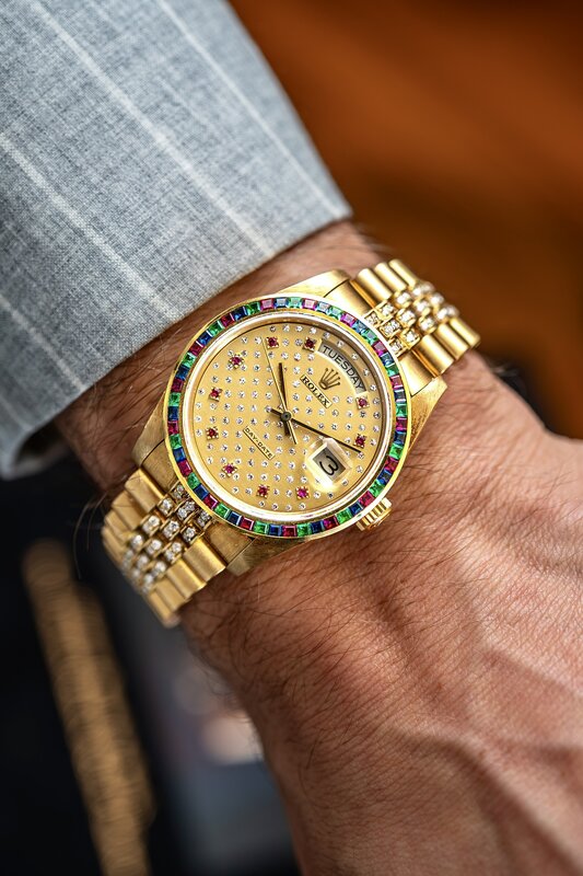 Same rainbow-bezel Day-Date from another angle — gold case, pavé dial, light grey suit sleeve