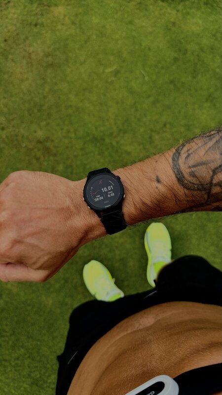 A Garmin watch on the wrist reading 10.01 km at 6:48 per kilometre, neon shoes on grass below