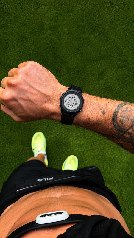 A Garmin watch on the wrist over turf reading 18.01 km, heart rate 157, neon shoes below