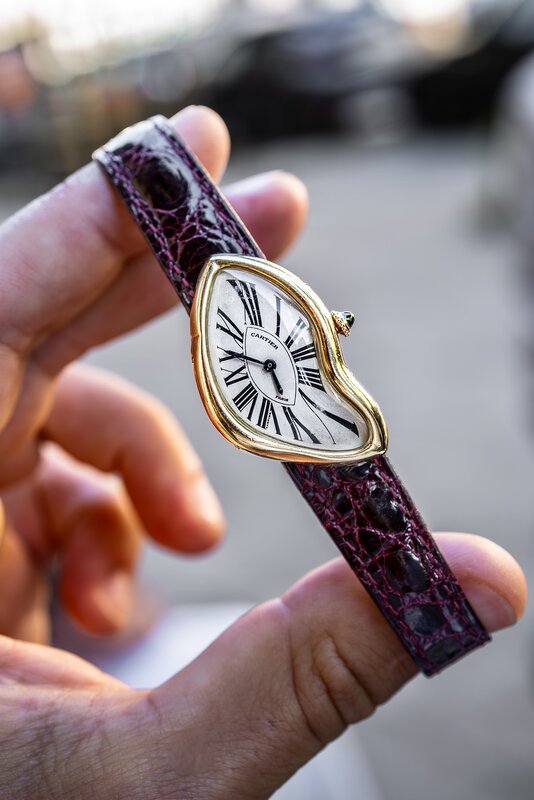 Yellow-gold Cartier Crash held between finger and thumb, its warped case and stretched Roman numerals on a burgundy alligator strap
