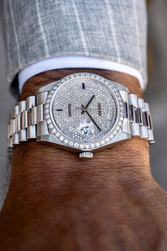 A Rolex Day-Date with a pavé-set dial and diamond-set bezel on a white-metal bracelet, worn beneath a grey pinstripe cuff