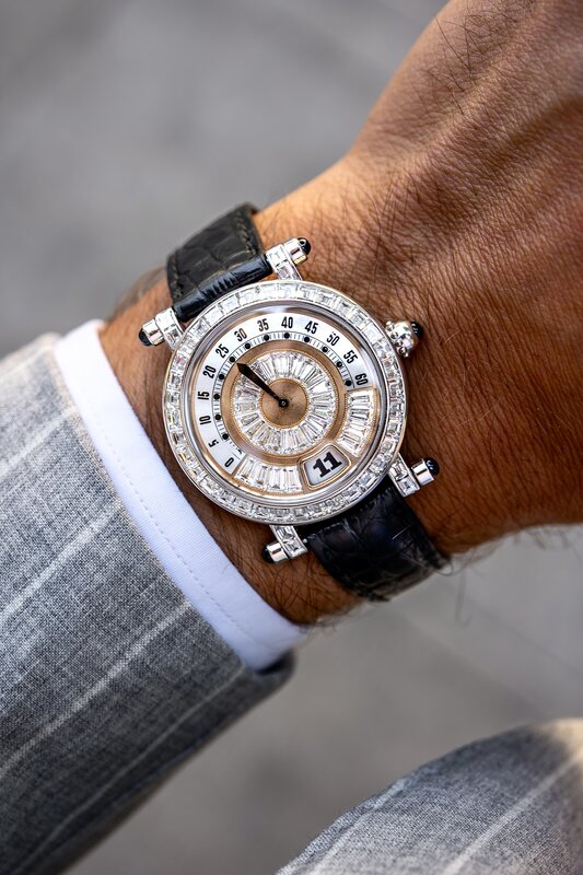 A watch with concentric rings of baguette-cut clear stones around its dial, on a black strap beneath a grey suit cuff