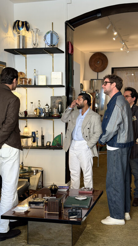 Four men standing around a low table in a small room lined with framed art, shelves and decorative objects