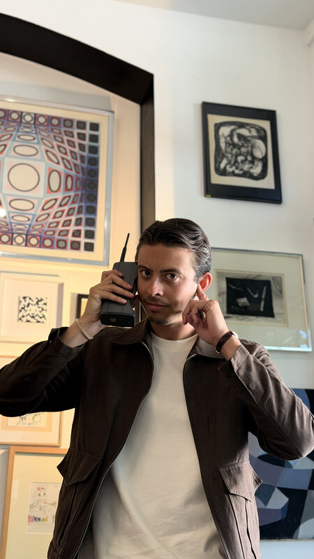 A man in a brown jacket holding a large vintage object with a short antenna to his ear, with framed prints covering the wall behind him