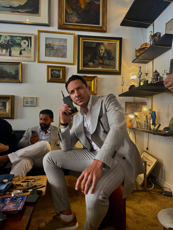 A man in a grey pinstripe suit holding a large vintage object with a short antenna to his ear in a room filled with framed art and display shelves