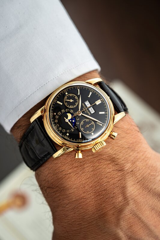 A yellow-gold Patek Philippe perpetual calendar chronograph with a black dial and moon phase, on a black alligator strap under a white shirt cuff