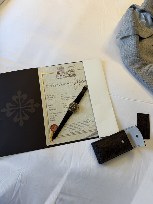 A black-dial Patek Philippe 2499 resting on its Extract from the Archives document on a white hotel bed, beside a black folder and leather pouch