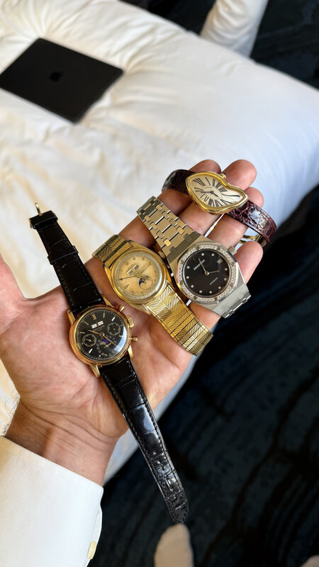 A hand holding four watches at once, gold and steel together, over a white hotel bed with a laptop beside