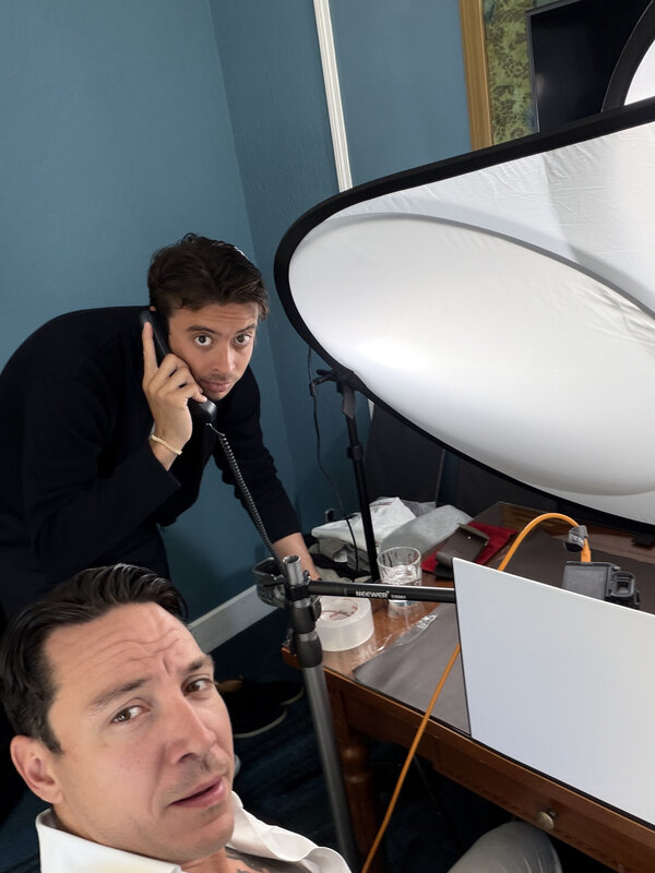 A hotel-room shoot in progress, a large softbox over the table, Jimmy in the selfie foreground and someone on the phone behind