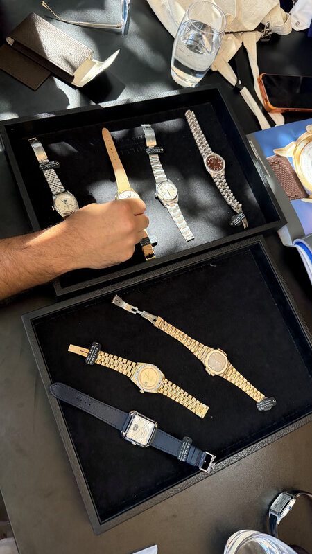 A hand reaching over an open display tray of watches at a viewing, more trays laid out along the table