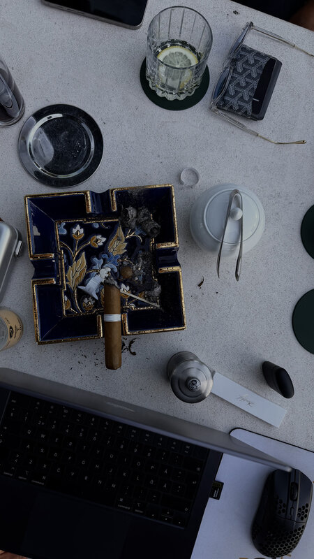 Overhead view of a table with a cigar in an ornate ashtray, two glasses, part of a laptop keyboard and several small personal items