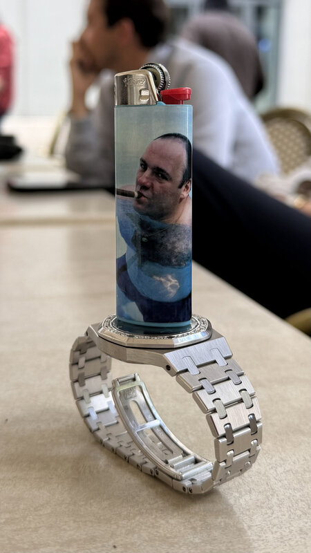A disposable lighter with a printed portrait standing upright on the white-gold bracelet of a diamond-bezel Audemars Piguet, a 750 hallmark on the clasp, on a marble table