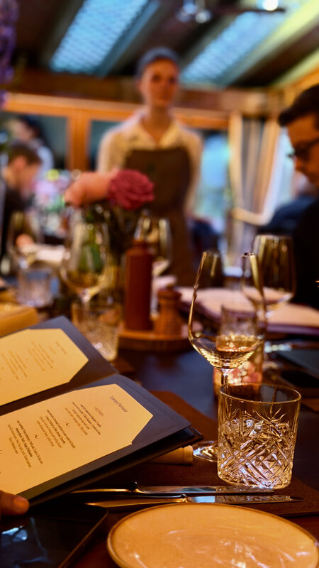 A long restaurant table set for dinner, menus, flowers and cut-glass tumblers, the room busy behind