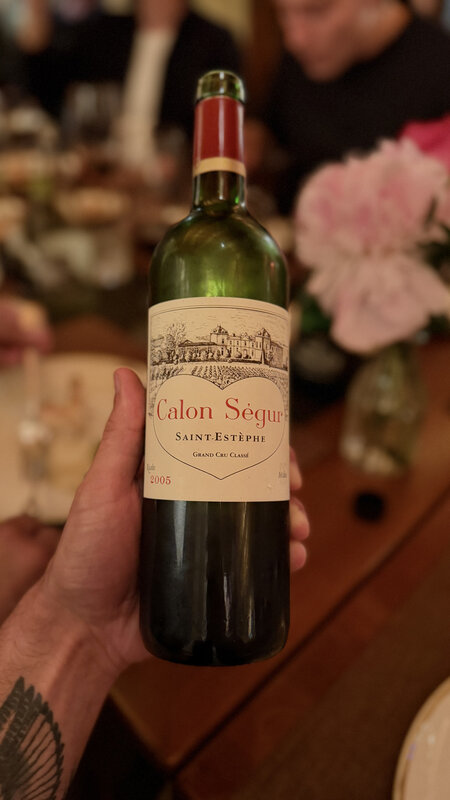 A hand holding a bottle of Château Calon Ségur 2005, Saint-Estèphe, above the dinner table