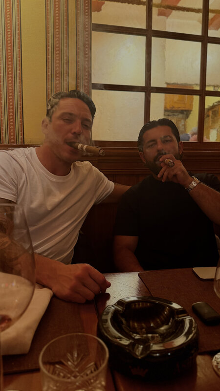 Two men with cigars at a restaurant table, tall windows behind them, an ashtray and glasses on the table