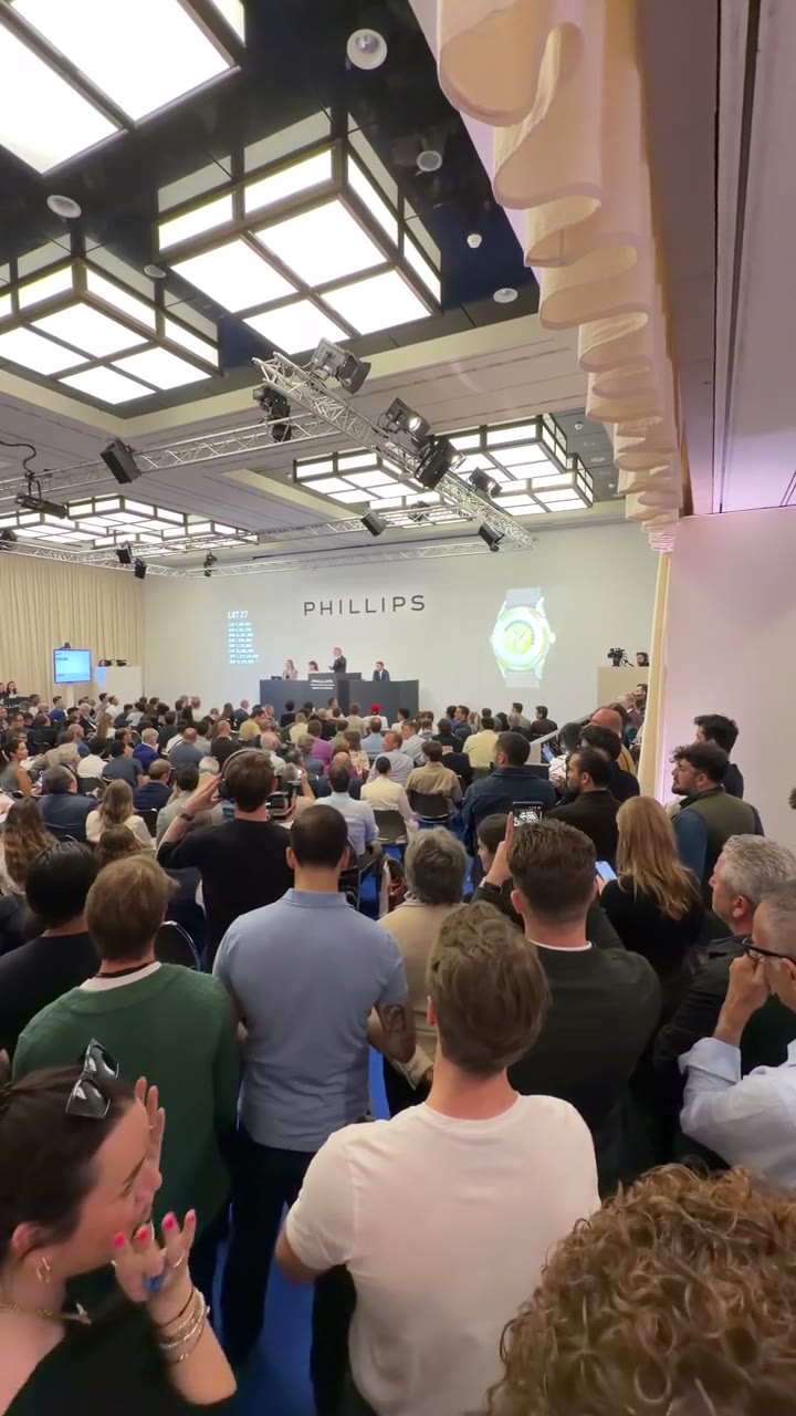 Video: the camera moves through a crowded Phillips saleroom towards the auctioneer at the rostrum while a watch is displayed on the projection screen