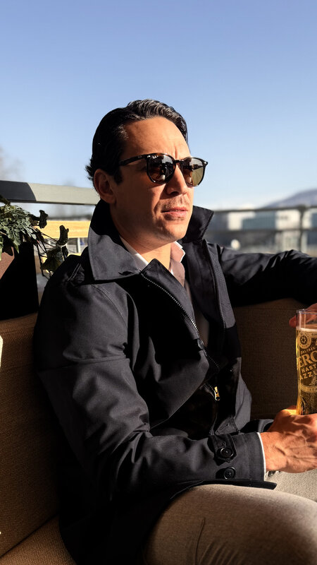 Jimmy in dark sunglasses and a navy trench coat on a Geneva terrace, holding a drink, soft afternoon light
