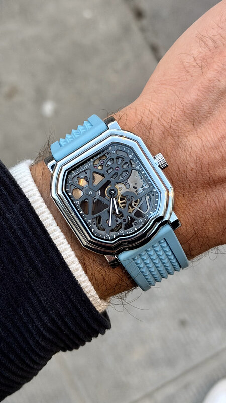 Independent maker watch with skeleton dial and light blue rubber strap on wrist, black ribbed sweater sleeve