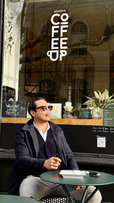 Jimmy seated outside Coffee Up Genève on a sunny afternoon — sunglasses, dark blazer