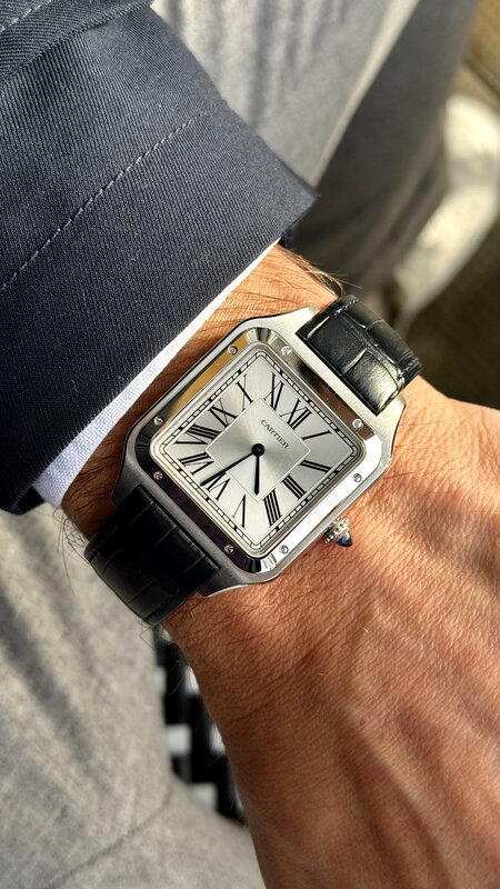 Cartier Santos on wrist with black leather strap, grey suit sleeve, soft daylight