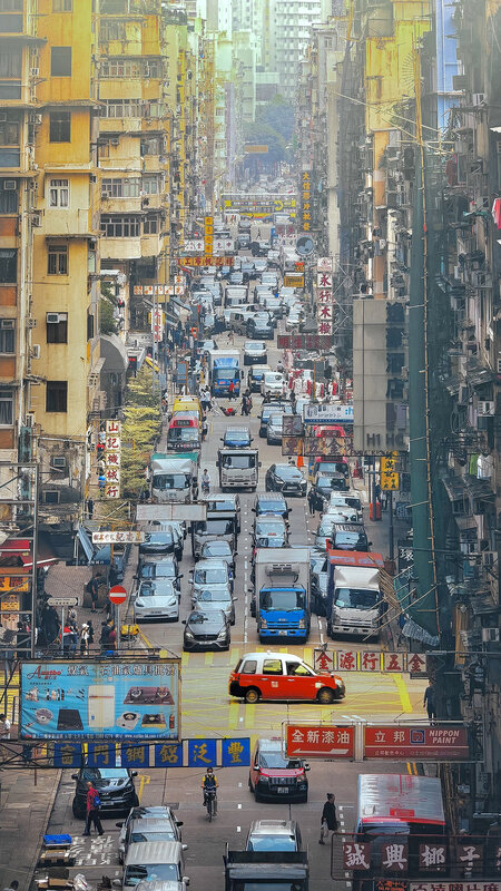 Dense Hong Kong street seen from above, packed traffic, red taxis and Chinese shop signs between tall residential blocks