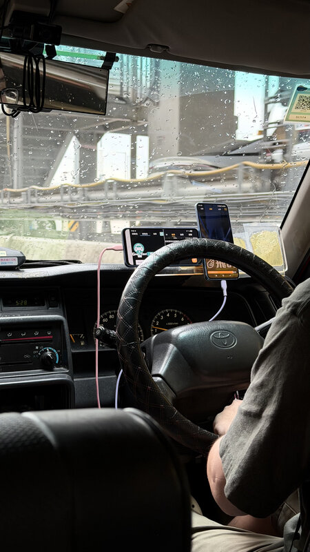 View from the back seat of a Hong Kong taxi, a braided wheel and the driver's arm resting low, a wet windscreen and the city beyond
