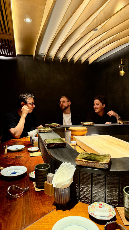 A stone counter set with serving slabs under a curved wooden slat ceiling, three people seated behind it