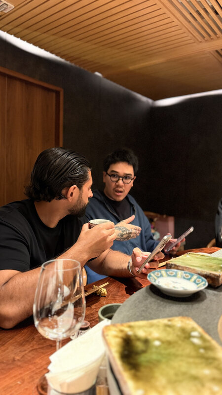 Two men at a restaurant table in Hong Kong, wine glasses and small plates between them, wood-slat ceiling above