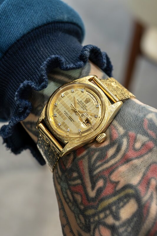 Yellow-gold Rolex with a textured gold dial and diamond hour markers, on a heavily tattooed wrist, blue knit cuff pushed back